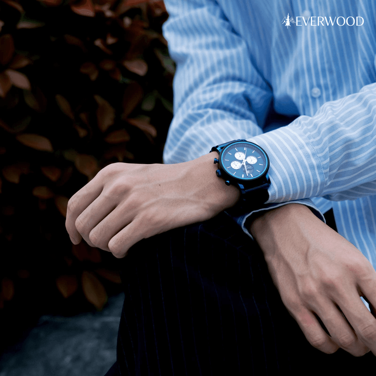 Férfi fa karóra kék számlappal, csíkos ingben, kültéri háttérrel.